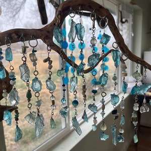 Puede incluir: Un carillón de madera flotante con múltiples hilos de cuentas de vidrio azul y transparente y cristales que cuelgan de él. El carillón está colgado frente a una ventana.