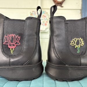 以下が含まれることがあります： 黒いアンクルブーツのペア。両サイドに伸縮性のあるパネルが付いています。各ブーツには、ピンクと黄色の蓮の花の刺繍が施されています。ブーツの後ろにはプルタブがあり、厚い黒いソールが付いています。