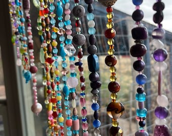 May include: A colorful beaded curtain with strands of various colors, including blue, red, green, and purple. The beads are made of glass, wood, and other materials. The curtain is hanging in front of a window.