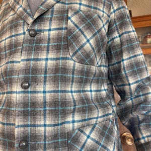 May include: A gray, blue, and white plaid jacket with three dark gray buttons and a chest pocket. The jacket has a classic collar and a tailored fit, suitable for cooler weather. The plaid pattern is a mix of gray, white, and blue lines.