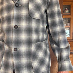 May include: A gray and cream plaid jacket with three dark brown buttons and two front pockets. The jacket has long sleeves with buttoned cuffs. The fabric has a soft, textured appearance.