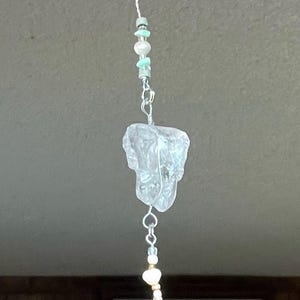 May include: A teal, blue, and white glass and crystal beaded suncatcher with a silver chain and a silver ring. The suncatcher is hanging from a ceiling.