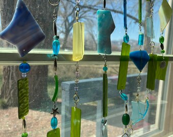 Könnte beinhalten: Ein Fenster-Sonnenfänger mit verschiedenen blauen und grünen Glasscheiben, Perlen und Metallakzente. Der Sonnenfänger hängt an einem Fenster mit Blick auf Bäume und einen strahlend blauen Himmel.