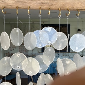 Puede incluir: Una cortina de conchas blancas con múltiples conchas redondas colgando de una viga de madera. Las conchas están enhebradas con un hilo delgado y cuelgan en un patrón escalonado.