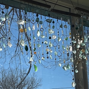 May include: A window with a handmade glass and metal wind chime hanging in front of it. The chime is made of various shades of blue and green glass beads and metal rings. The wind chime is hanging from a wooden dowel rod.