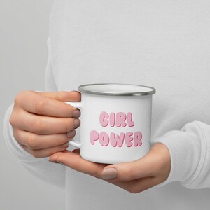 Può includere: Tazza smaltata bianca con bordo e manico argentati. La tazza presenta le parole "GIRL POWER" in lettere rosa a bolle. La tazza è tenuta tra le mani di una persona.