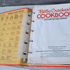 May include: A vintage Betty Crocker's Cookbook, new and revised edition, with a red and white cover and a yellow spine. The cookbook is open to a page with a chart of herbs and spices. The cookbook has a ring binder and includes several orange index tabs.