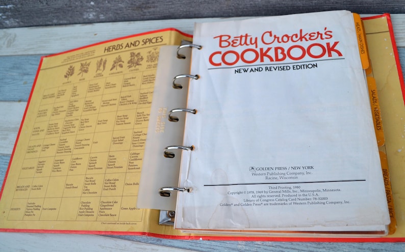 May include: A vintage Betty Crocker's Cookbook, new and revised edition, with a red and white cover and a yellow spine. The cookbook is open to a page with a chart of herbs and spices. The cookbook has a ring binder and includes several orange index tabs.