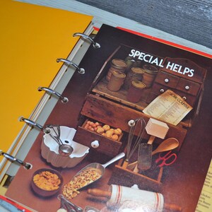 May include: A yellow binder with a red and white cookbook inside. The cookbook has the title "Special Helps" and a section titled "Cook Course". The cookbook is open to a page with a picture of a kitchen cabinet with various cooking utensils and ingredients.