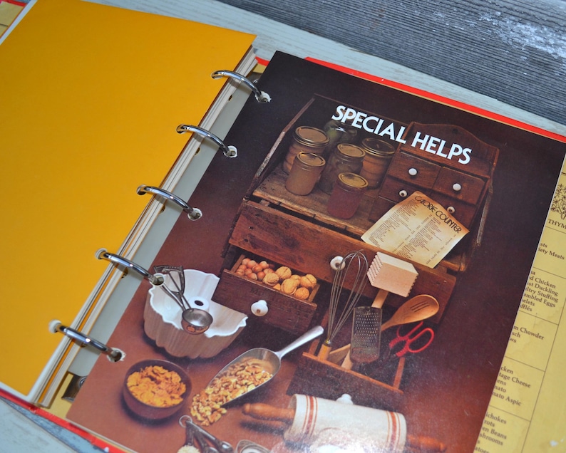 May include: A yellow binder with a red and white cookbook inside. The cookbook has the title "Special Helps" and a section titled "Cook Course". The cookbook is open to a page with a picture of a kitchen cabinet with various cooking utensils and ingredients.