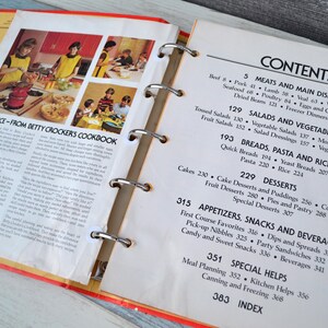 May include: A close-up of an open cookbook with a red and yellow cover. The book is titled "Confidence - From Betty Crocker's Cookbook". The pages are filled with recipes and a table of contents listing different categories of recipes, including meats, salads, breads, desserts, appetisers, snacks, beverages, and special helps.
