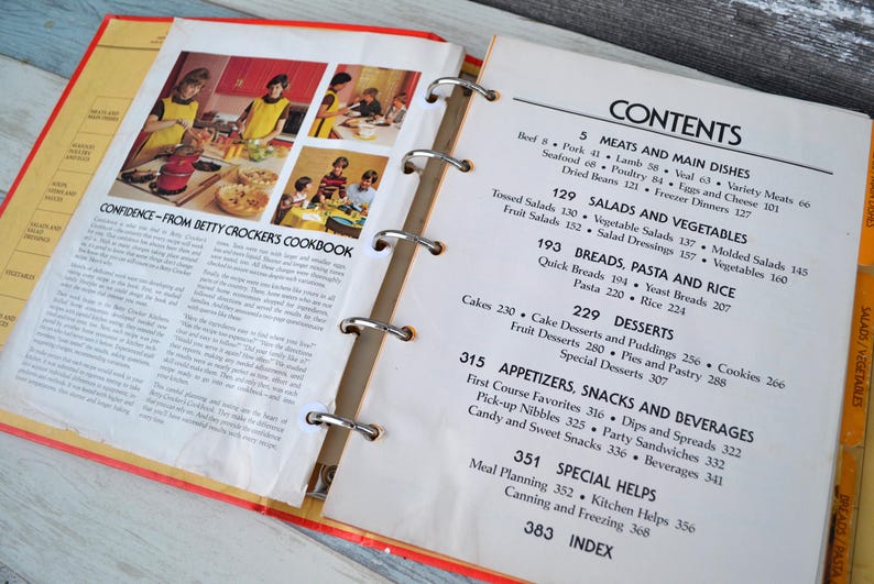May include: A close-up of an open cookbook with a red and yellow cover. The book is titled "Confidence - From Betty Crocker's Cookbook". The pages are filled with recipes and a table of contents listing different categories of recipes, including meats, salads, breads, desserts, appetisers, snacks, beverages, and special helps.
