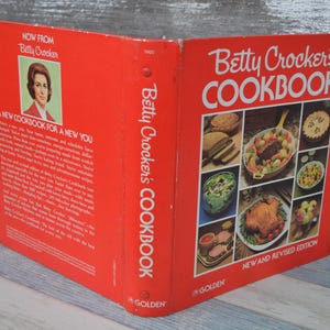 May include: A red and white Betty Crocker's Cookbook featuring a new and revised edition. The cover showcases a selection of food photographs, including a casserole, chicken, and salad.