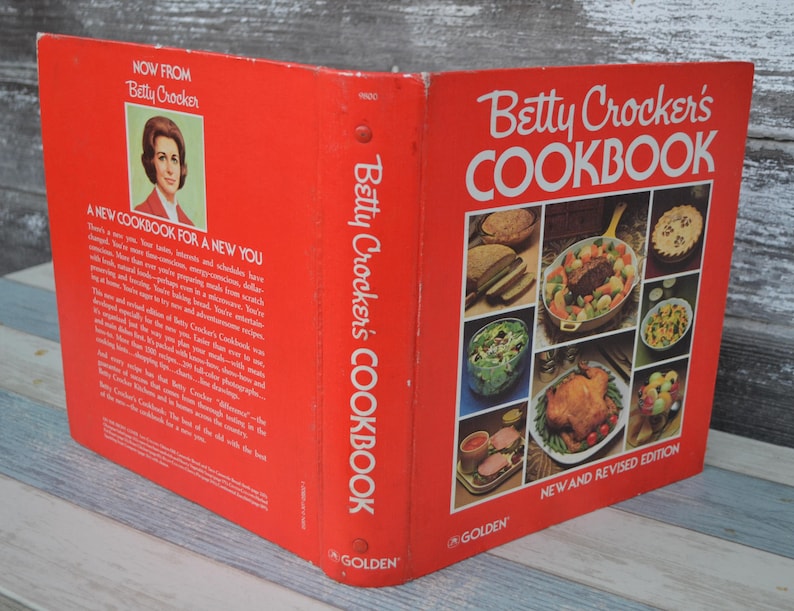 May include: A red and white Betty Crocker's Cookbook featuring a new and revised edition. The cover showcases a selection of food photographs, including a casserole, chicken, and salad.