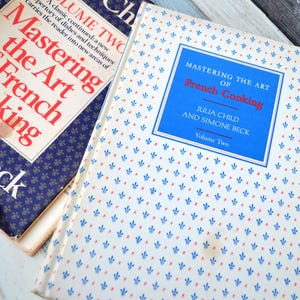 May include: Two vintage cookbooks, "Mastering the Art of French Cooking," are displayed. The books have blue and white covers with red text. One book has a floral pattern, and the other has a dark blue background with white text.
