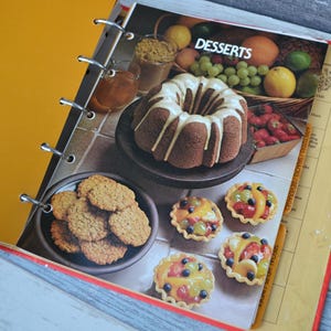 May include: A binder with a yellow cover and a page open to a recipe section titled "Desserts". The page features a chocolate cake with white frosting, a plate of cookies, and several mini fruit tarts.