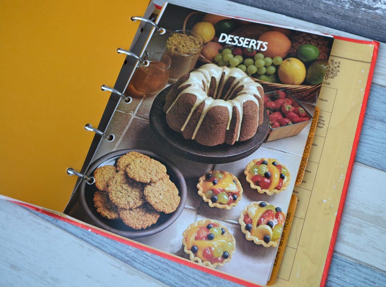 May include: A binder with a yellow cover and a page open to a recipe section titled "Desserts". The page features a chocolate cake with white frosting, a plate of cookies, and several mini fruit tarts.