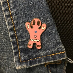 May include: Pink gingerbread person enamel pin with the word "F.U." on the arms. The pin is on a blue denim jacket.
