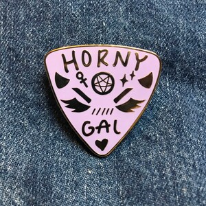 Könnte beinhalten: Eine violette, dreieckige Emaille-Anstecknadel mit goldener Umrandung. Die Anstecknadel zeigt den Text "Horny Gal" mit einem Pentagramm, Flügeln und anderen Symbolen in Schwarz.