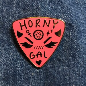 May include: Pink enamel pin with a gold outline. The pin is shaped like a triangle with rounded corners. It says "Horny Gal" in black lettering with a pentagram, an ankh, and wings in the center. There is a small heart at the bottom.