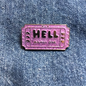 May include: A pink glitter enamel pin shaped like a ticket with the word "HELL" in black letters and the text "ADMIT ONE" in smaller black letters. The numbers "666" and "999" are on either side of the ticket.