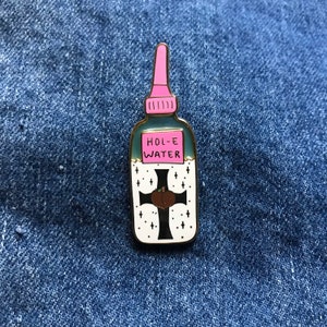 May include: A gold-colored enamel pin shaped like a bottle of holy water. The bottle is blue and white with a pink dropper. The label reads "HOL-E WATER" and features a black cross with two peach halves on it.