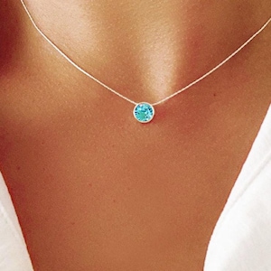 Op de afbeelding: Een delicate zilveren ketting met een enkele, ronde, lichtblauwe edelsteen hanger. De hanger hangt aan een dunne zilveren ketting. De ketting wordt getoond tegen een wit overhemd.