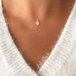 May include: A gold oval pendant featuring a small embossed figure, suspended on a thin, clear nylon cord necklace. The pendant rests against the skin, and the text Leslucilles is written in white cursive script in the lower right corner.