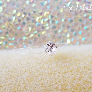 May include: A delicate, clear ring with a single, sparkling, round-cut, white gemstone. The ring band is nearly invisible, creating the illusion that the gem floats. The background is a soft, textured surface with a shimmering, iridescent effect.