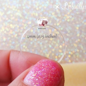 May include: A clear ring with a small, square, clear gem. The ring is 4mm (0.15 inches) in diameter. The ring is held by a person with pink glitter nail polish. The background is a bokeh of white and gold.