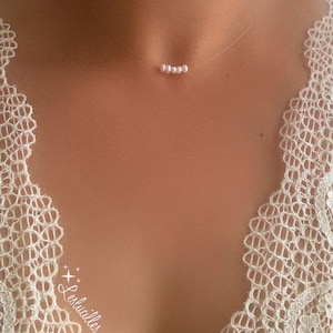 Peut inclure: Un collier délicat composé de cinq petites perles blanches montées sur un fil transparent. Le collier est porté sur la poitrine d'une personne, avec un vêtement en dentelle blanche visible sur le côté gauche. L'ensemble est minimaliste et élégant.