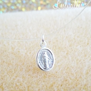 May include: A silver oval pendant necklace featuring an image of the Virgin Mary. The pendant is suspended on a clear, nearly invisible cord. The pendant has the words "ORIGINALLY CONCEIVED O.R.A. 1930" around the edge.