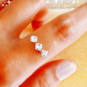 May include: Three square-cut diamond studs are displayed on a finger. The diamonds are clear and sparkling, set in a minimalist design. The skin tone is light, and the background is a soft, blurred gold.