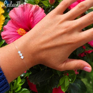 Könnte beinhalten: Ein zartes Armband mit drei kleinen, klaren, rund facettierten Steinen in silberfarbener Fassung, befestigt an einer dünnen, transparenten Nylonschnur, die das Handgelenk umschließt. Das Design ist schlicht und hebt die Steine hervor.