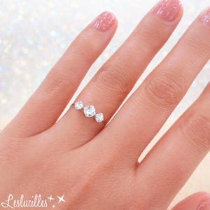 May include: A silver ring with three clear, square-cut stones. The ring is on a finger with pink nail polish. The ring is a simple design, with the stones set close together. The ring is likely made of sterling silver or white gold.