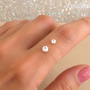 Op de afbeelding: Een ketting van transparant nylondraad met twee kleine, ronde, briljant geslepen witte stenen in zilverkleurige zettingen. De stenen zijn op verschillende hoogtes aan het koord bevestigd, wat een zwevend effect op de huid creëert.