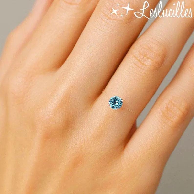 May include: A delicate ring featuring a round, light blue gemstone. The ring appears to be held by a nearly invisible band, creating a floating effect. The image includes the text "Leslucilles" in the upper right corner.