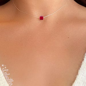 May include: A delicate necklace featuring a small, ruby-red square bead. The bead is suspended on a thin, silver-colored chain. The necklace is worn close to the neck, creating a minimalist and elegant look.