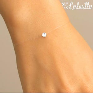 Peut inclure: Un bracelet minimaliste avec un seul diamant rond et transparent sur un fil presque invisible. Le bracelet est porté sur un poignet. Le nom de la marque "Leslucilles" est visible.