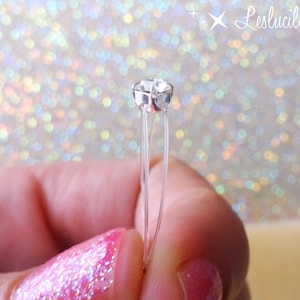 May include: A clear, faceted crystal bead on thin, transparent wires. The bead is held between fingers with pink nail polish. The background is a sparkly, iridescent bokeh effect. The text "Leslucilles" is in the upper right corner.