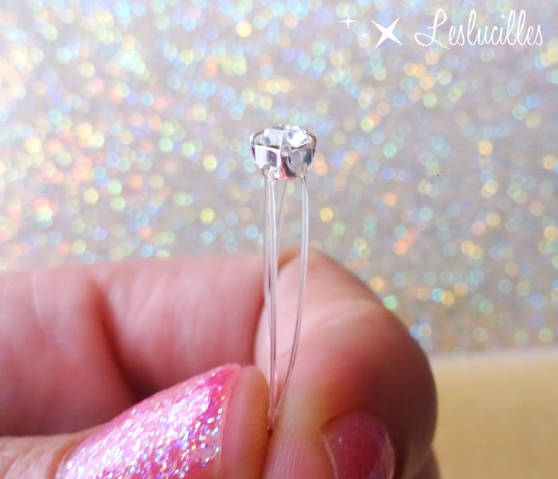 May include: A clear, faceted crystal bead on thin, transparent wires. The bead is held between fingers with pink nail polish. The background is a sparkly, iridescent bokeh effect. The text "Leslucilles" is in the upper right corner.