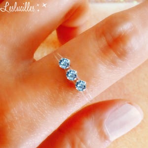 Peut inclure: Une bague délicate ornée de trois pierres bleues claires. Les pierres sont serties sur un fil transparent, créant un effet flottant. La bague est portée sur un doigt, avec la marque "Leslucilles" en haut à gauche.