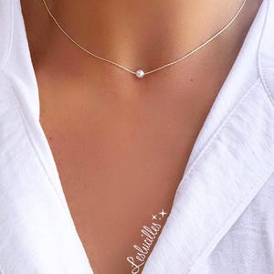 May include: Delicate gold-tone necklace featuring a single, small, round, white pearl pendant. The necklace is worn against a white shirt. The jewelry is simple and elegant.