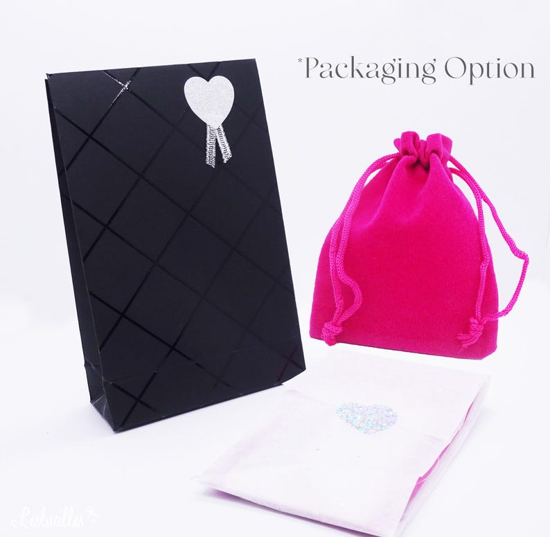 May include: An assortment of packaging options, including a black gift bag with a diamond pattern and a silver heart sticker, a bright pink velvet drawstring bag, and a white pouch with a heart sticker. The text "Packaging Option" is visible.