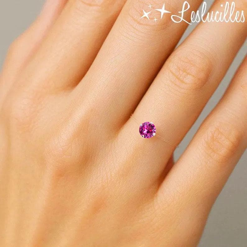 May include: A delicate ring featuring a vibrant, round, magenta gemstone. The ring appears to be held by a nearly invisible band, creating the illusion that the gem floats on the finger. The ring is set against a soft, neutral background.