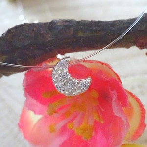 May include: A silver crescent moon pendant necklace with small clear crystals. The necklace is on a clear string and is hanging from a branch.