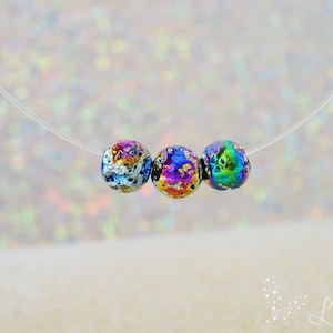 Può includere: Una collana con catena d'argento con un filo trasparente attaccato. Il filo ha tre perline iridescenti con un motivo maculato. Le perline sono un mix di blu, verde, viola e oro.