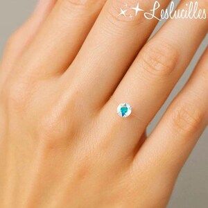 May include: A delicate ring featuring a clear band and a round, iridescent gemstone. The gemstone reflects shades of blue and green. The ring is worn on a finger, showcasing its minimalist design. The brand name "Leslucilles" is in the top right corner.