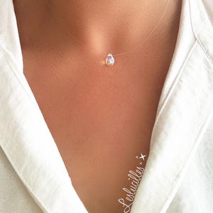 May include: A delicate necklace featuring a small, iridescent teardrop pendant suspended on a nearly invisible clear cord. The pendant reflects a spectrum of colors. The necklace is worn against a white shirt.