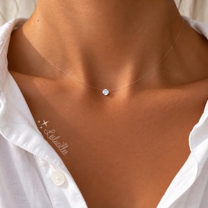 Peut inclure: Un collier en fil de nylon transparent avec un pendentif en pierre précieuse blanche ronde taille brillant centré à la base du cou. Le texte Leslucilles avec deux petites étoiles est imprimé en blanc sur la chemise blanche à col.